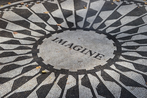 A circular mosaic pattern featuring geometric shapes in black and white, with the word 'IMAGINE' written at the center. The mosaic is situated outdoors and a few fallen leaves can be seen scattered across the surface.