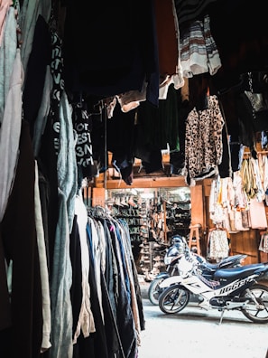 A marketplace stall filled with various clothing items hanging from above and arranged on racks along the sides. There are two motorcycles parked in the center of the area, underneath the hanging clothes. The stall appears to extend into a larger market with more hanging garments and accessories in the background. The lighting is a mix of natural and artificial, creating contrasting shadows and highlights.
