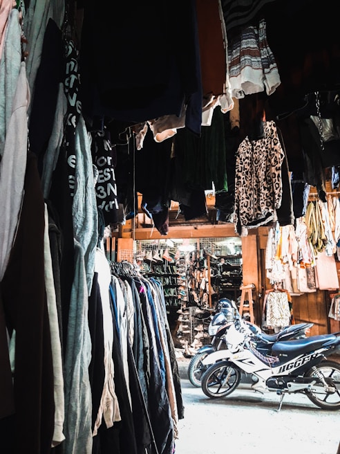 A marketplace stall filled with various clothing items hanging from above and arranged on racks along the sides. There are two motorcycles parked in the center of the area, underneath the hanging clothes. The stall appears to extend into a larger market with more hanging garments and accessories in the background. The lighting is a mix of natural and artificial, creating contrasting shadows and highlights.