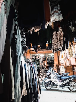 A marketplace stall filled with various clothing items hanging from above and arranged on racks along the sides. There are two motorcycles parked in the center of the area, underneath the hanging clothes. The stall appears to extend into a larger market with more hanging garments and accessories in the background. The lighting is a mix of natural and artificial, creating contrasting shadows and highlights.