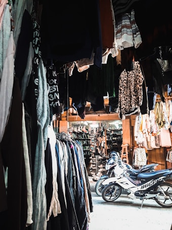 A marketplace stall filled with various clothing items hanging from above and arranged on racks along the sides. There are two motorcycles parked in the center of the area, underneath the hanging clothes. The stall appears to extend into a larger market with more hanging garments and accessories in the background. The lighting is a mix of natural and artificial, creating contrasting shadows and highlights.