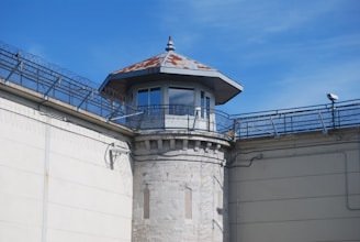 A guard tower with a rusted, pointed roof is positioned at the corner of a high stone wall. The wall is topped with barbed wire and the sky is a clear blue. Security cameras are attached to the walls, enhancing the sense of surveillance.