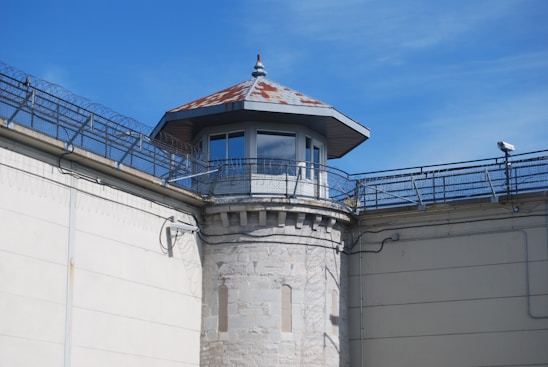 A guard tower with a rusted, pointed roof is positioned at the corner of a high stone wall. The wall is topped with barbed wire and the sky is a clear blue. Security cameras are attached to the walls, enhancing the sense of surveillance.