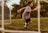 A young person wearing gloves and athletic clothing is in motion on a grassy soccer field, near a goalpost. The background features lush greenery, suggesting an outdoor sports setting. The lighting indicates a warm, possibly late afternoon or early evening scene.