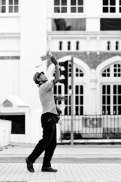 A person is engaged in juggling or performing with objects, captured in mid-action. The background features an urban setting with architecture that includes large windows and decorative arches.