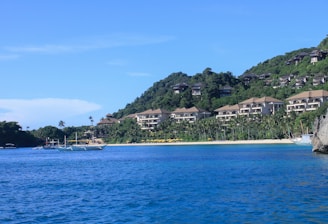 A serene coastline with clear blue waters extends to a sandy beach with lush greenery. Several boats are anchored near the shore. In the background, multiple resort-style buildings are nestled on a hillside covered with dense vegetation.