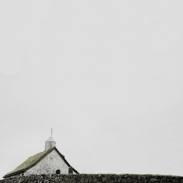 A small white church with a moss-covered roof and a cross on top stands behind an old stone wall. The sky is overcast, creating a serene and tranquil atmosphere.