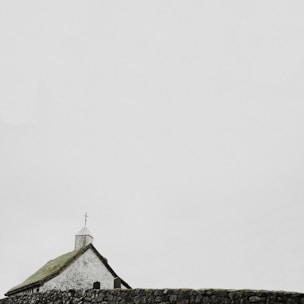 A small white church with a moss-covered roof and a cross on top stands behind an old stone wall. The sky is overcast, creating a serene and tranquil atmosphere.