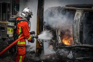 A firefighter in full gear is extinguishing a fire coming from a car that appears to have been in an accident and is overturned. The area is filled with thick smoke, and flames are visible. The surroundings look industrial, with signs and doors indicating a commercial or garage space nearby.