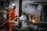 A firefighter in full gear is extinguishing a fire coming from a car that appears to have been in an accident and is overturned. The area is filled with thick smoke, and flames are visible. The surroundings look industrial, with signs and doors indicating a commercial or garage space nearby.