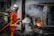A firefighter in full gear is extinguishing a fire coming from a car that appears to have been in an accident and is overturned. The area is filled with thick smoke, and flames are visible. The surroundings look industrial, with signs and doors indicating a commercial or garage space nearby.