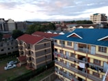Several apartment buildings are visible in a residential area. The forefront features a multi-story building with blue and red roofs and clothes hanging on the balconies. Additional buildings and a few trees are seen in the background beneath a cloudy sky.