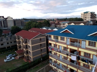 Several apartment buildings are visible in a residential area. The forefront features a multi-story building with blue and red roofs and clothes hanging on the balconies. Additional buildings and a few trees are seen in the background beneath a cloudy sky.