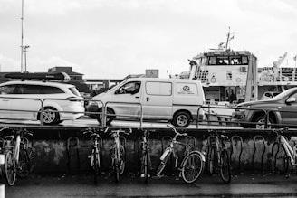 A monochrome photograph depicts a harbor scene with several parked vehicles, including a van and a car with a roof box. In the background, a large ferry or ship is visible, moored alongside. In the foreground, there is a row of bicycles secured to a railing, with wet pavement indicating recent rain.