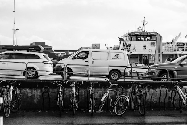 A monochrome photograph depicts a harbor scene with several parked vehicles, including a van and a car with a roof box. In the background, a large ferry or ship is visible, moored alongside. In the foreground, there is a row of bicycles secured to a railing, with wet pavement indicating recent rain.