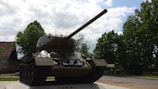 A military tank is positioned on a platform during a partly cloudy day. Surrounding the tank are lush green trees, and a road runs alongside the scene. The tank, an armored vehicle, is situated prominently in the foreground, emphasizing its long barrel and heavy-duty tracks.