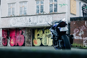 A motorcycle is parked in front of a building with graffiti-covered walls. The walls display a variety of colorful graffiti art, including the text 'OLGA & PIOTR FAST MOOD' and other vibrant designs. The building has several windows, some of which are boarded up. The scene suggests an urban environment with an artistic and edgy atmosphere.