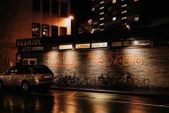 A nighttime street scene featuring a building with illuminated signage reading 'Paahjul' and bicycle-related designs on the exterior wall. Several bicycles are parked outside the building, and a car is parked on the roadside. The street is wet, possibly from recent rain, reflecting the warm lights from the building and nearby streetlights.