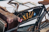 A vintage bicycle with a worn appearance features a brown leather saddle and an attached old-fashioned suitcase or tool box. The bicycle frame has the letters 'NBSDI' painted in yellow, with a small Norwegian flag and another emblem nearby. The image conveys a sense of nostalgia and rustic charm.