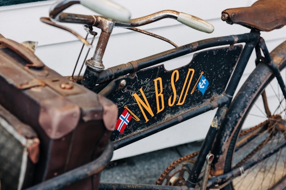 A vintage bicycle with a worn appearance features a brown leather saddle and an attached old-fashioned suitcase or tool box. The bicycle frame has the letters 'NBSDI' painted in yellow, with a small Norwegian flag and another emblem nearby. The image conveys a sense of nostalgia and rustic charm.