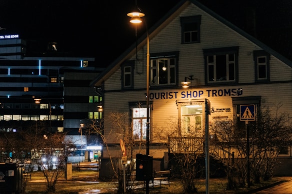 A nighttime street scene featuring a wooden building with a sign that reads 'Tourist Shop Troms&oslash;'. The building is illuminated by warm street lamps, and there are bare trees in the foreground. Nearby, a modern building with horizontal stripes and lit windows is visible.