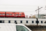 A red train with visible branding and symbols is stationary on the railway tracks. Below it, another train with a grey and black design passes by. A white van is partially visible in the foreground. Overhead, electric lines are stretched across the sky. A couple of people walk on a platform, and there are advertisements and signage nearby. The environment appears industrial and utilitarian.