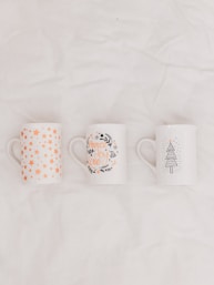 three white-and-multicolored ceramic mugs on white textile
