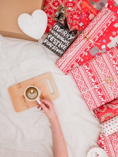 A person holds a cup with latte art on a wooden board next to a collection of wrapped Christmas presents. The gifts have red and white festive wrapping paper with designs such as reindeer and snowflakes, arranged on a light surface. A white heart-shaped object with the word 'Faith' and a blackboard sign with Christmas greetings are also visible.