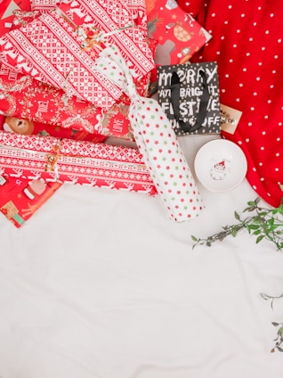 A collection of wrapped presents in festive red and white wrapping paper with patterns featuring snowflakes and stars. One package is in a bottle shape, wrapped with white paper adorned with red and green stars. There is also a small bag with the words 'Merry and Bright' in black and white. A small dish with a festive design is placed beside the gifts. Greenery is partially visible on the side, adding a touch of nature.