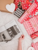 A hand holding open a black and white photo book lying on a white sheet, surrounded by several brightly wrapped Christmas gifts in red and white patterns. A wooden heart with the word 'Faith' is visible, along with a black sign with festive text. The scene conveys a cozy and festive atmosphere.