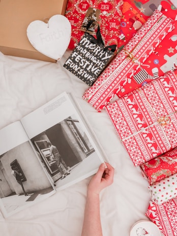 A hand holding open a black and white photo book lying on a white sheet, surrounded by several brightly wrapped Christmas gifts in red and white patterns. A wooden heart with the word 'Faith' is visible, along with a black sign with festive text. The scene conveys a cozy and festive atmosphere.