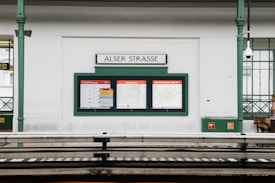 A train station platform featuring signage for 'Alser Strasse' with informational displays. The platform has a green and white color scheme, with a waiting area and security camera visible. Architectural elements include decorative columns and frosted glass windows.