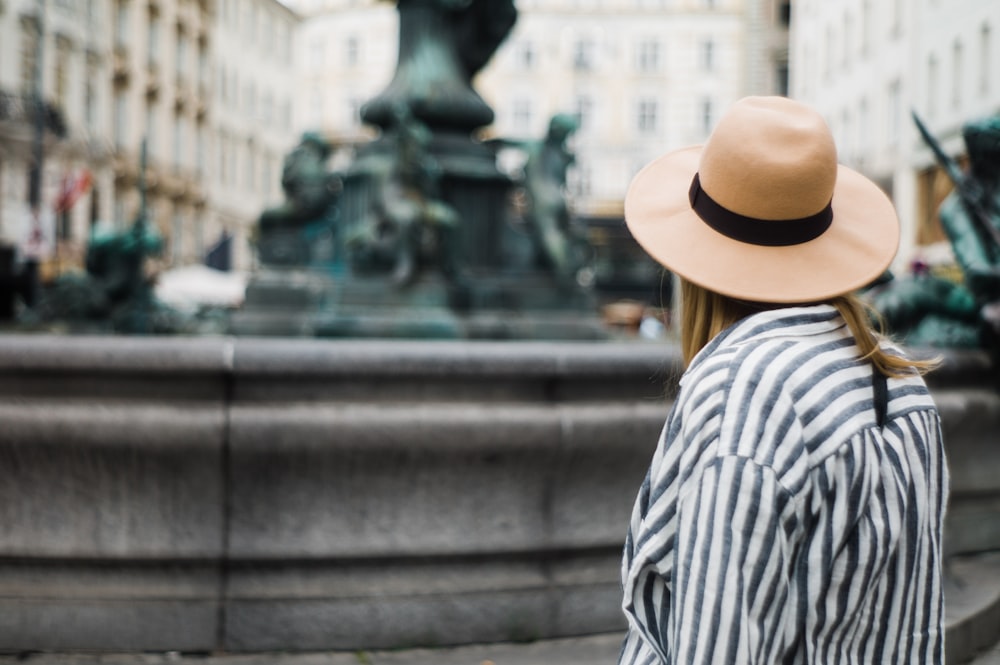 woman wearing gray and white striped shirt and brown hat while standing near gray fountain during daytime