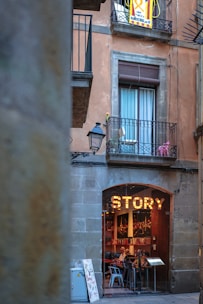 A rustic building facade with a large sign reading 'STORY' above the entrance of a cozy, inviting cafe or restaurant. The establishment is visible through an arched doorway, where several patrons are seated at tables inside. Above, a balcony with a window is adorned with some potted plants and a banner with text, suggesting a local or cultural significance. The setting conveys a quaint and picturesque urban setting.