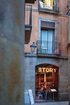 A rustic building facade with a large sign reading 'STORY' above the entrance of a cozy, inviting cafe or restaurant. The establishment is visible through an arched doorway, where several patrons are seated at tables inside. Above, a balcony with a window is adorned with some potted plants and a banner with text, suggesting a local or cultural significance. The setting conveys a quaint and picturesque urban setting.