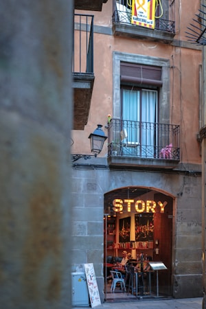 A rustic building facade with a large sign reading 'STORY' above the entrance of a cozy, inviting cafe or restaurant. The establishment is visible through an arched doorway, where several patrons are seated at tables inside. Above, a balcony with a window is adorned with some potted plants and a banner with text, suggesting a local or cultural significance. The setting conveys a quaint and picturesque urban setting.
