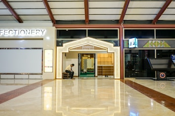 A well-lit entrance to a prayer room with a decorative arch is located in a spacious area with a reflective floor. A person is seated on a bench near the entrance, which is flanked by a confectionery shop and another establishment with a geometric sign.