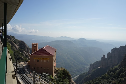 A mountainous landscape with a prominent view of an orange-roofed building labeled 'Montserrat' perched on the edge of a steep hill. The scene includes a railway track leading away from the building. The background reveals layers of distant mountains under a clear blue sky with scattered clouds.