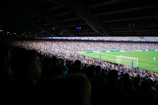 A large crowd fills the stands of a stadium, watching a soccer match on a brightly lit field. The spectators are packed closely together, creating a sea of varied colors and textures. Advertising banners display prominently around the field.