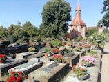 A well-maintained cemetery featuring stone graves adorned with a variety of colorful flowers. In the background, a large tree provides shade, and an ornate chapel with a red roof and spire adds a sense of tranquility. The surrounding environment is lush with greenery.