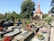 A well-maintained cemetery featuring stone graves adorned with a variety of colorful flowers. In the background, a large tree provides shade, and an ornate chapel with a red roof and spire adds a sense of tranquility. The surrounding environment is lush with greenery.
