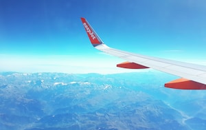 An airplane wing with an easyJet logo is visible, flying over a vast mountain range with snow-capped peaks. The clear sky is a bright blue, hinting at a sunny day, and the expansive view showcases various shades of blue and white.