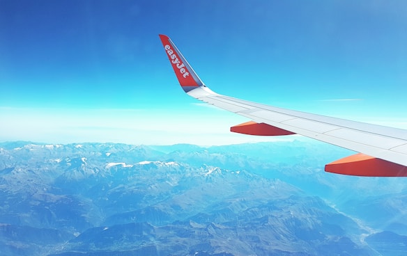 An airplane wing with an easyJet logo is visible, flying over a vast mountain range with snow-capped peaks. The clear sky is a bright blue, hinting at a sunny day, and the expansive view showcases various shades of blue and white.