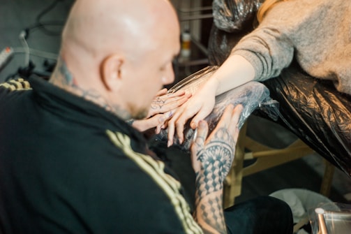 A person with tattooed hands is working on another person's hand, which is wrapped for protection. The setting has a focused and artistic atmosphere, likely in a tattoo studio.