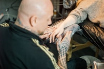 A person with tattooed hands is working on another person's hand, which is wrapped for protection. The setting has a focused and artistic atmosphere, likely in a tattoo studio.