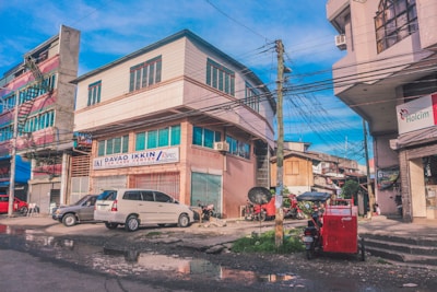 A street scene with a large building marked 'Davao Ikkin Car Care Center.' The surrounding area includes other smaller buildings, power lines, and a variety of parked vehicles such as cars and motorcycles. The street appears wet with puddles, indicating recent rain. Bright colors are present in the architecture and vehicles, creating a vibrant urban atmosphere.