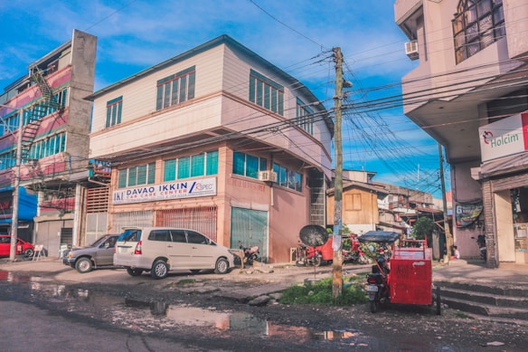A street scene with a large building marked 'Davao Ikkin Car Care Center.' The surrounding area includes other smaller buildings, power lines, and a variety of parked vehicles such as cars and motorcycles. The street appears wet with puddles, indicating recent rain. Bright colors are present in the architecture and vehicles, creating a vibrant urban atmosphere.