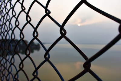 A chain-link fence in the foreground with a blurred background that includes a body of water and a distant horizon. The sun is peeking through a cloudy sky, creating a serene atmosphere.