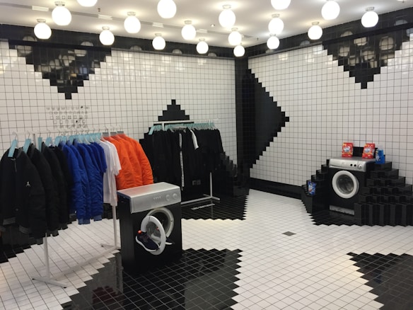 A stylish room designed with black and white tiled walls and floor, featuring geometric patterns. Clothing items in black, blue, orange, and white hang on racks to the left. Two washing machines are placed in the room, with one having detergent boxes on top.