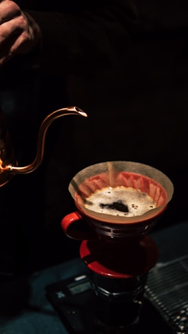 A person is pouring water from a copper gooseneck kettle into a red pour-over coffee dripper with freshly brewed coffee. The scene is dimly lit, highlighting the textured surface of the dripper and the bubbling coffee. The setup includes a scale beneath the dripper.